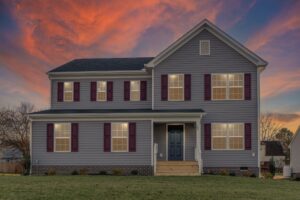 A two-story house with gray siding, a dark front door, and red shutters, lights on inside, stands before a dramatic sunset with orange and purple clouds in the sky.