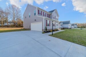 Two-story house with light gray siding and red shutters, featuring a spacious driveway, front garage, small porch, and well-maintained front lawn, all under a clear sky with some clouds.