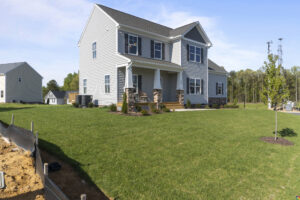 A two-story house with white siding and a gray roof, featuring a front porch with columns, placed on a large grass lawn with minimal landscaping and an unfinished area near the left.