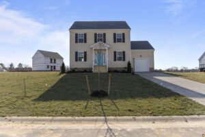 A tan two-story house with black shutters and a single-car garage is situated on a freshly seeded lawn. A newly planted tree is in front. A partially constructed house is next door.