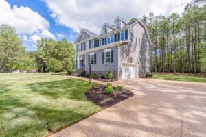 A two-story gray house with white trim, blue shutters, and manicured landscaping sits on a large grassy lot next to a forested area, under a partly cloudy sky.