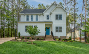 A two-story house with a blue front door and gray roof, surrounded by a well-maintained lawn and trees in the background. The driveway is on the left side.
