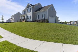 A modern two-story house with a large green lawn and a clear blue sky, viewed from a curving sidewalk.