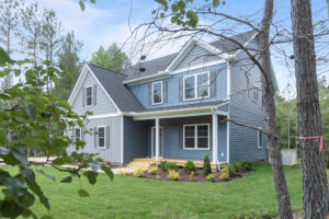 A two-story blue house with white trim and a covered porch, surrounded by a green lawn and trees, under a clear sky.