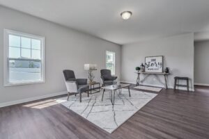 Modern living room with two gray armchairs, a coffee table on a patterned rug, a floor lamp, and a decorative sign reading "Our story begins here." Two windows provide natural light.