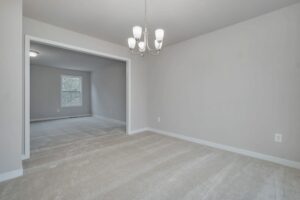 Empty room with beige carpeted flooring, a white ceiling, and light gray walls. A chandelier hangs from the ceiling. Another room with a window is visible through the open doorway.