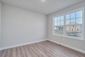 A small empty room with light-colored walls and wood flooring. A large window on one wall provides a view of a suburban neighborhood.