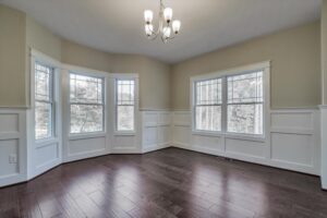 An empty room with dark hardwood floors, beige walls, white wainscoting, a chandelier, and multiple large windows providing natural light.