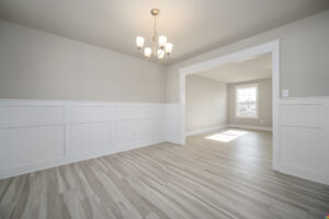 A bright, empty room with light wood floors, white wainscoting, and a chandelier. An open doorway leads to another sunlit room with a large window.