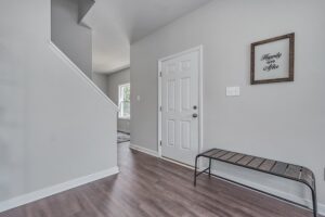 Interior of a home featuring a white door, a wooden bench, grey walls, a framed "happiness here" sign, and a staircase.