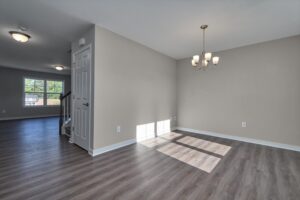A vacant room with light gray walls, dark wood flooring, and a chandelier. A closed door is seen on the left side, leading to a stairway, and a large window is visible in the background.