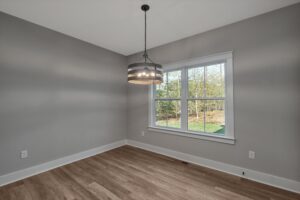 A vacant room with gray walls, a large window with a view of trees, a modern hanging light fixture, and wooden flooring.