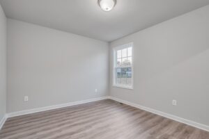 Empty, light gray room with wood flooring, a single ceiling light fixture, and a window on the right wall.
