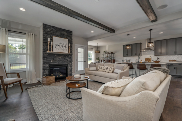 A modern living room with a stone fireplace, beige sofas, a wooden coffee table, and a kitchen in the background. The room features wooden beams on the ceiling and a large rug on the floor.