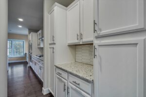 A modern kitchen with white cabinets, granite countertops, stainless steel appliances, and a hexagon tile backsplash. The room has dark wood floors and a view of a window at the end of the hallway.
