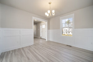 A bright, empty room with light gray wood flooring, white wainscoting walls, a window, and a chandelier. An open doorway leads to a hallway with a front door and additional windows.