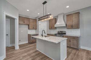 A modern kitchen with light wood cabinets, a white island with a built-in sink, stainless steel appliances, including a stove with a hood, and a central pendant light fixture. The floor is wood.