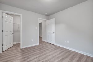A vacant room with light gray walls, light-colored hardwood flooring, a closed door, an open door leading to a closet, and another open door leading to a hallway.
