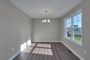 An empty room with light gray walls, wood flooring, a central ceiling light fixture, and a large window letting in natural light.