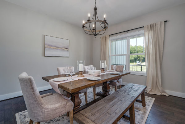 A dining room with a wooden table and bench, upholstered chairs, a chandelier, a framed artwork on the wall, and a large window with beige curtains. The table is set for a meal.