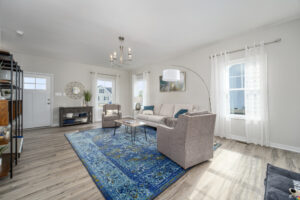 Bright, modern living room with two beige sofas, a blue rug, and minimalistic decor under a chandelier, featuring large windows with sheer curtains.