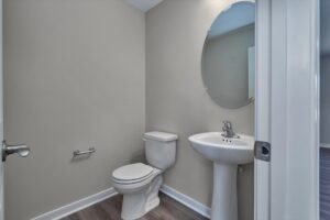 A small bathroom with a toilet, pedestal sink, and oval mirror on the wall. The floor has wood-like tiles, and the walls are painted light gray.