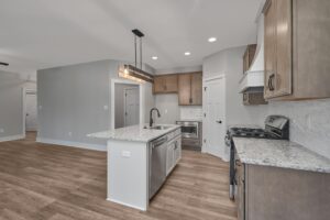 A modern kitchen with light wood cabinetry, granite countertops, an island with a sink, a gas stove, stainless steel appliances, and hardwood floors. The kitchen is well-lit with recessed and pendant lights.
