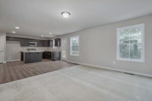 An empty open-concept living area with a view of the kitchen. The room has light-colored walls, a mix of carpet and hardwood flooring, and large windows letting in natural light.