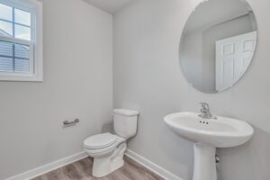 A clean, modern bathroom features a white toilet, a pedestal sink, and a round mirror on the wall. A window brings in natural light, and the wood floor complements the neutral wall color.
