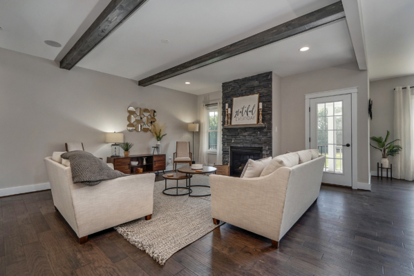 A modern living room with beige sofas, a stone fireplace, wooden beams on the ceiling, and a glass door leading outside. The room has a neutral color scheme and contemporary decor.