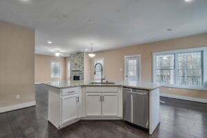A modern kitchen with a central island, granite countertops, a built-in dishwasher, and white cabinetry. The open floor plan extends to the living area with large windows and a stone fireplace.