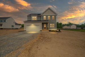 A two-story house with an illuminated garage and front porch, set against a sunset sky. The surroundings are mostly bare ground with sparse greenery. Adjacent houses are visible on either side.