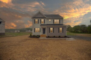 A two-story house with a gray and white exterior stands on a dirt lot, with a cloudy sky at sunset in the background.
