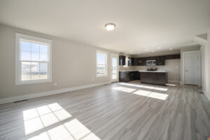 A spacious, unfurnished room with light wood flooring, large windows, and an adjacent modern kitchen featuring dark cabinetry and stainless steel appliances.