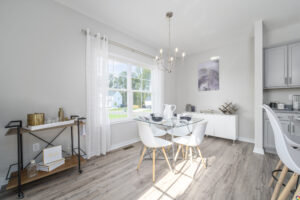 Modern dining room with a white table, four chairs, and serving cart, featuring large windows with sheer curtains and a minimalist chandelier.