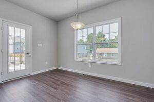 Empty room with gray walls, dark wooden floor, and a large window showing a suburban view, next to an open door.