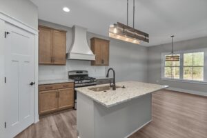 Modern kitchen with light wood cabinets, a white range hood, stainless steel appliances, and an island with a sink and granite countertops. Light fixture above the island, window with natural light.