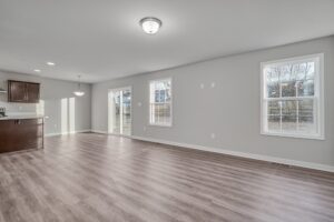 A spacious, unfurnished open-plan living area with light grey walls and wooden flooring, featuring ample natural light from large windows and sliding glass doors leading to an outdoor space.