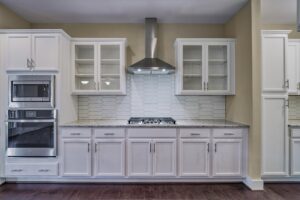 A modern kitchen features white cabinets, a hexagonal tile backsplash, stainless steel oven, microwave, and range hood, and a gas cooktop set in a granite countertop.