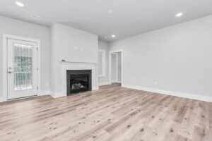 Spacious, empty living room with light hardwood floors, white walls, a fireplace, and a glass door leading outside.