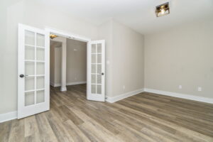 Empty room with beige walls, laminated wood flooring, and two open white french doors leading to other rooms.
