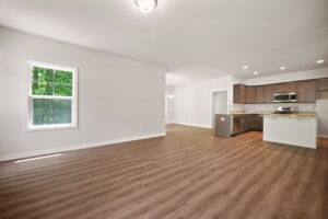 An empty, modern kitchen with stainless steel appliances and wooden cabinets, extending into a spacious living area with laminate flooring and large windows.