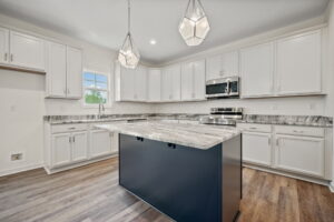 Modern kitchen with white cabinets, gray countertops, stainless steel appliances, and wooden flooring. Features a dark island with a marble top and geometric pendant lights hanging above.