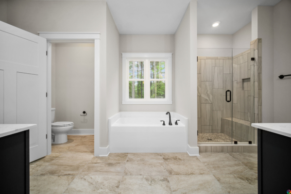 A modern bathroom with a bathtub, shower with glass door, toilet, and double sink vanity. The room has beige tiles and a window above the bathtub.