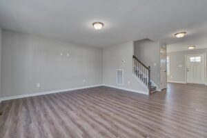 An empty room with wooden flooring, white walls, ceiling lights, and a staircase leading upstairs near a white door with a small window.