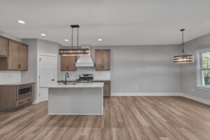 A modern kitchen with wooden cabinets, a central island with a sink, stainless steel appliances, two pendant lights, and light wood flooring. The adjacent area appears to be an empty dining space.