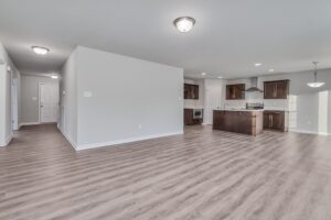 Spacious, empty open-plan living area with light gray walls, wood-look flooring, and a kitchen featuring dark brown cabinets and stainless steel appliances. A hallway leads to a door on the left.