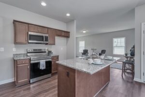 Modern kitchen with stainless steel appliances, granite countertops, and wooden cabinets. An island with dishes and a visible open living area with seating in the background.