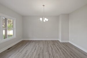 A vacant room with light gray walls, a window, a hanging light fixture, and hardwood flooring.