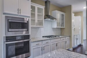 Modern kitchen with stainless steel appliances, including a microwave and oven, white cabinetry, a gas cooktop with a hood, and white tile backsplash. Granite countertops on island and cabinets.